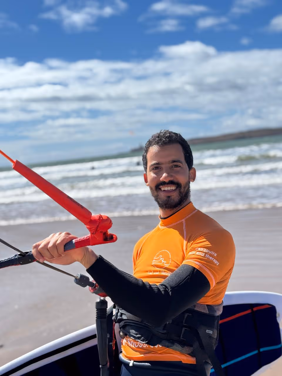 Sif Eddine on the Essaouira beach with surf and kite gear, ocean behind him